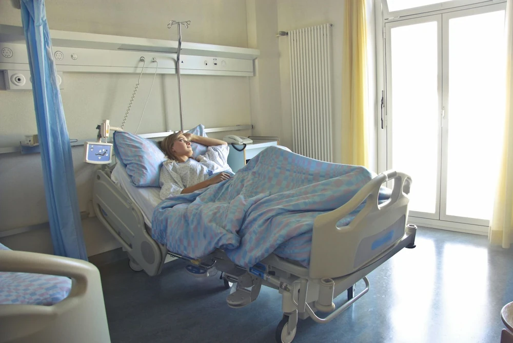 A young woman reclines in a hospital bed and looks out the glass door to her right.