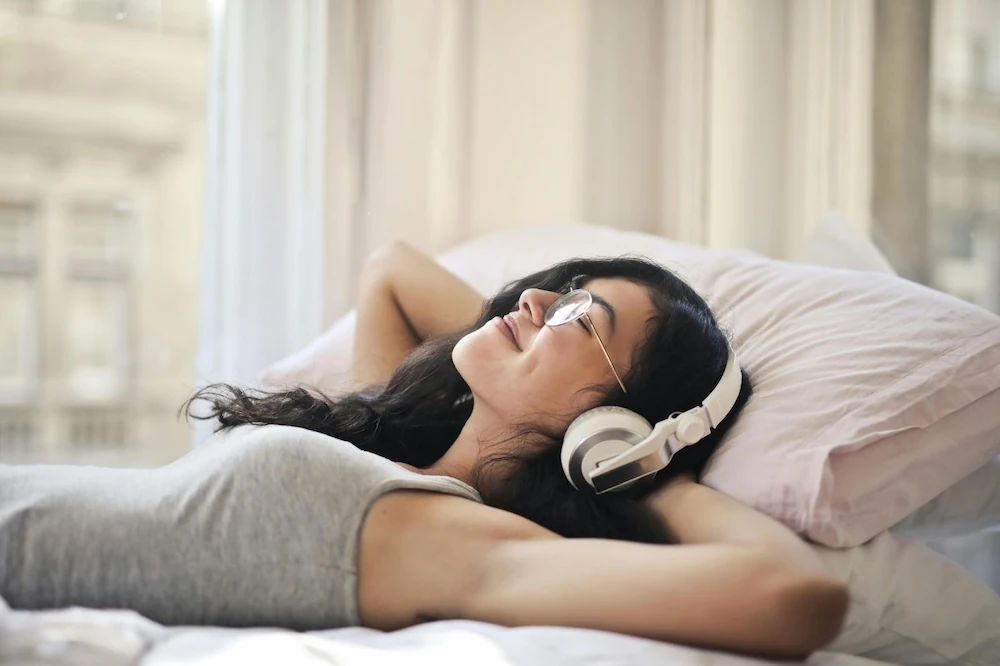 A woman in a gray tank top and wire-framed glasses wears white headphones and leans back on two pillows. Her hair is dark and she is smiling with her eyes closed.