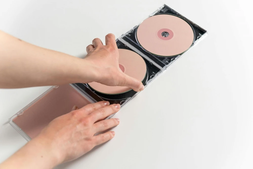 Two pink CDs, darker at the center, sit in CD cases next to each other. A hand reaches out to remote the left CD from its case.