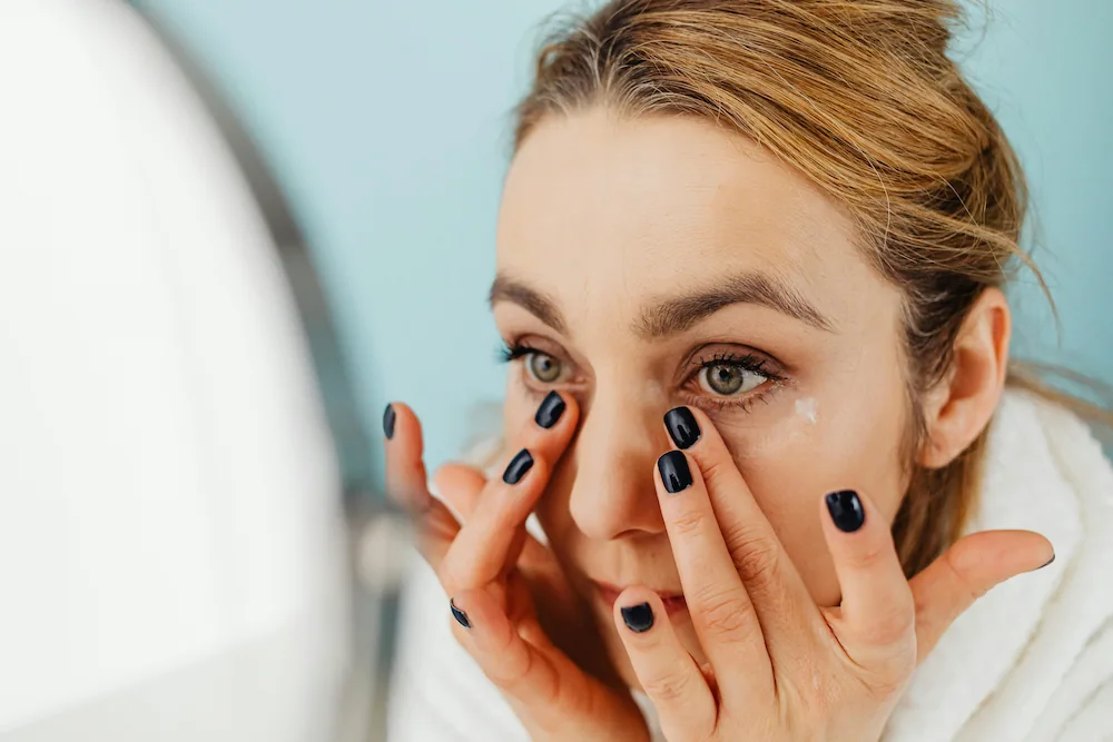 A woman wearing nail polish and eye makeup rubs the skin underneath her eyes while looking in a mirror.