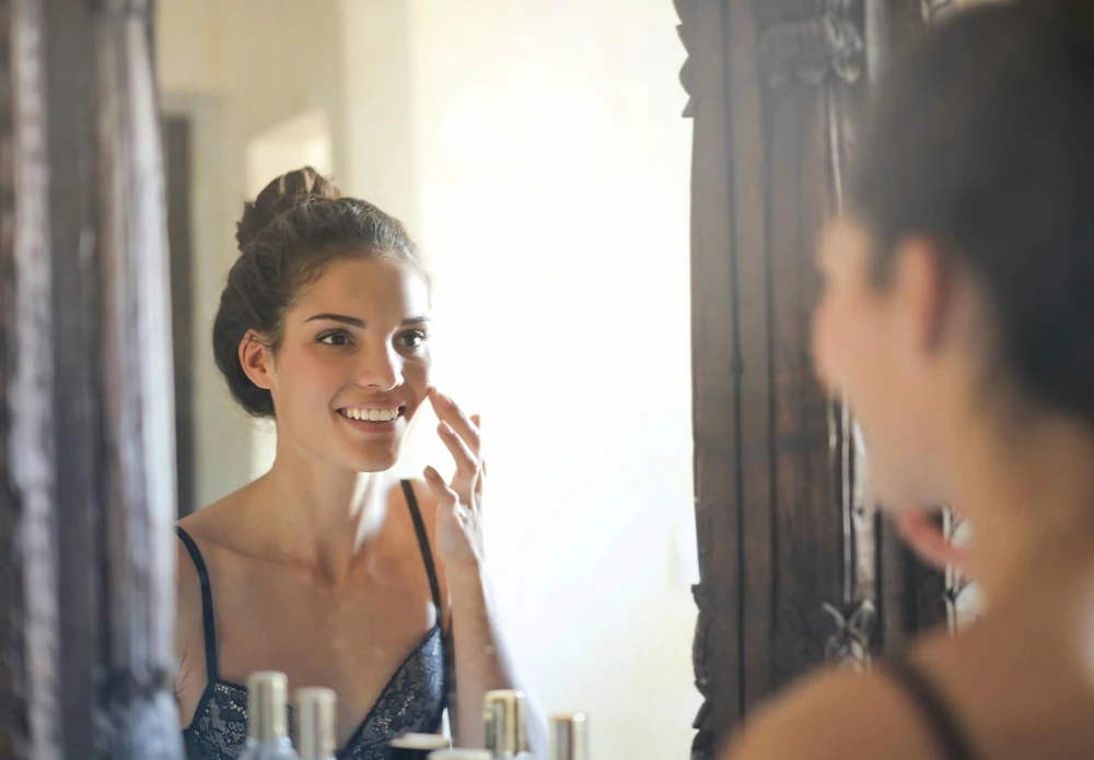 A woman with a bun in her hair smiles while rubbing her face, looking in the mirror.