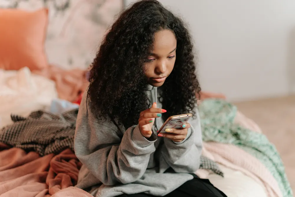 A young girl sits on her bed while looking at a smart phone.